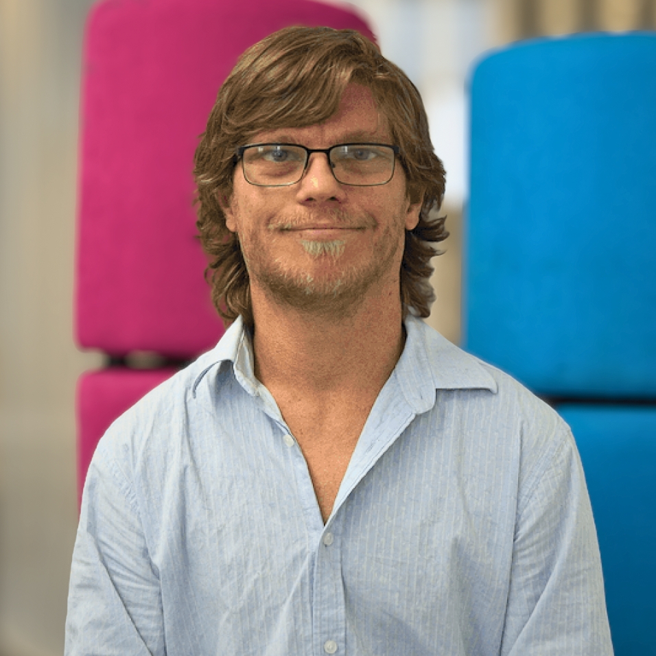 A man with wavy brown hair, glasses, and a short beard smiles slightly while looking at the camera. He is wearing a light blue button-up shirt. The background features large, colourful blocks in bright pink and blue tones.