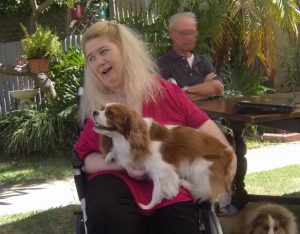 Photo of Ann Marie in a wheelchair with a king charles spaniel on her lap. She has long blonde hair and is wearing a red short sleeved top. There is a blurred man in the background.