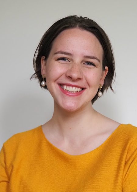Photo of Tessa smiling at camera. She has short, brown hair as is wearing an orange blouse, and has a light grey background behind her.