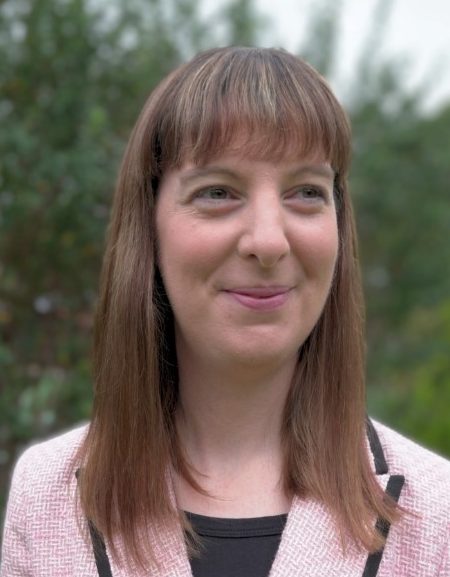A woman with straight, shoulder-length brown hair and fringe smiles softly while looking slightly to the side. She is wearing a light pink jacket with black trim over a black top. The background is an outdoor setting with blurred greenery.