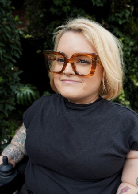 A woman with chin-length blonde hair smiles gently while looking off to the side. She is wearing oversized tortoiseshell glasses and a black t-shirt, with tattoos visible on both arms. Her hand rests on the control of a power wheelchair. There is blurred greenery behind her.