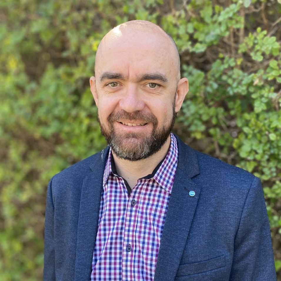 Photo of Brad, who is smiling at camera. He is bald and has a beard, and is wearing a navy blazer with a checked shirt. There is blurred greenery in the background.