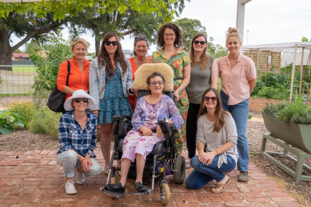A group of nine women pose together in an outdoor garden setting. One woman is seated in a power wheelchair, and the others are standing or crouching beside her, all smiling at the camera. The background includes raised garden beds and leafy trees.