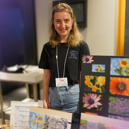 Erin stands, smiling at the camera wearing a black top and a lanyard. Photos and artwork in front of her.