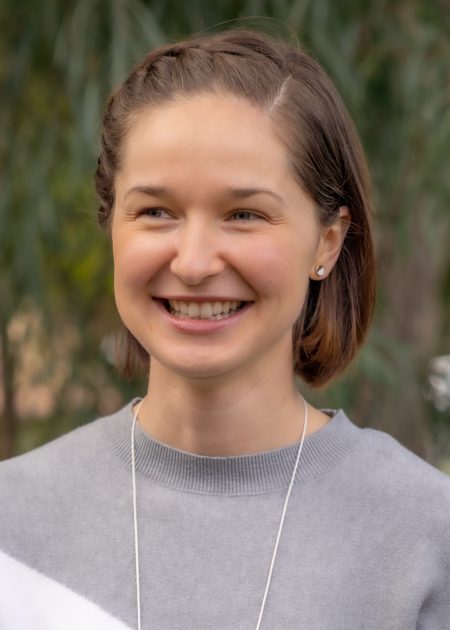 A woman with short, brown hair smiles while standing outdoors. She is wearing a grey and white color-blocked sweater. The background features blurred greenery.