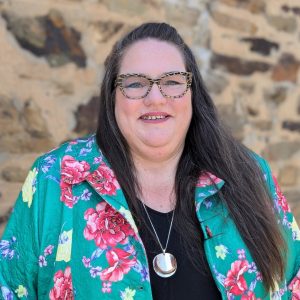 Heidi, a woman with long dark hair wearing dark framed glasses, is smiling at the camera. She is standing in front of a stone wall and is wearing a green floral jacket over a black top.