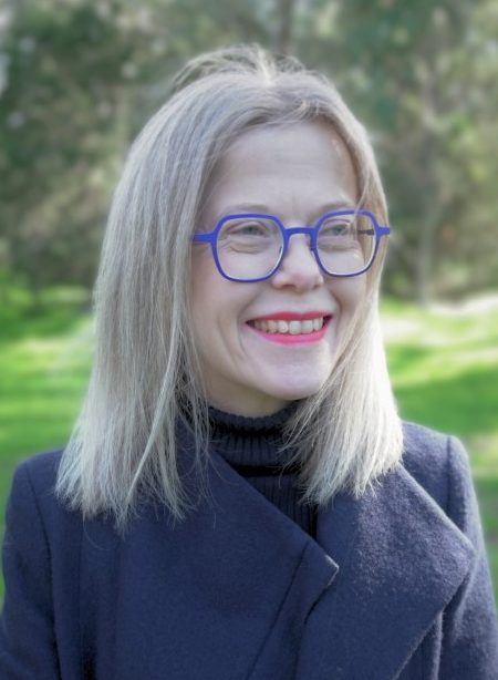 A woman with straight, shoulder-length blonde hair smiles while standing outdoors in a sunlit park. She is wearing bold blue glasses, bright pink lipstick, a black turtleneck, and a dark coat. The background is filled with greenery and softly blurred trees.