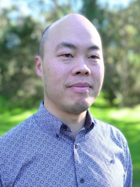 A man with a shaved head stands outdoors, looking slightly to the side with a calm expression. He is wearing a patterned button-up shirt. The background features soft-focus greenery and trees in natural sunlight.