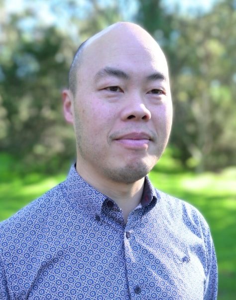 A man with a shaved head stands outdoors, looking slightly to the side with a calm expression. He is wearing a patterned button-up shirt. The background features soft-focus greenery and trees in natural sunlight.