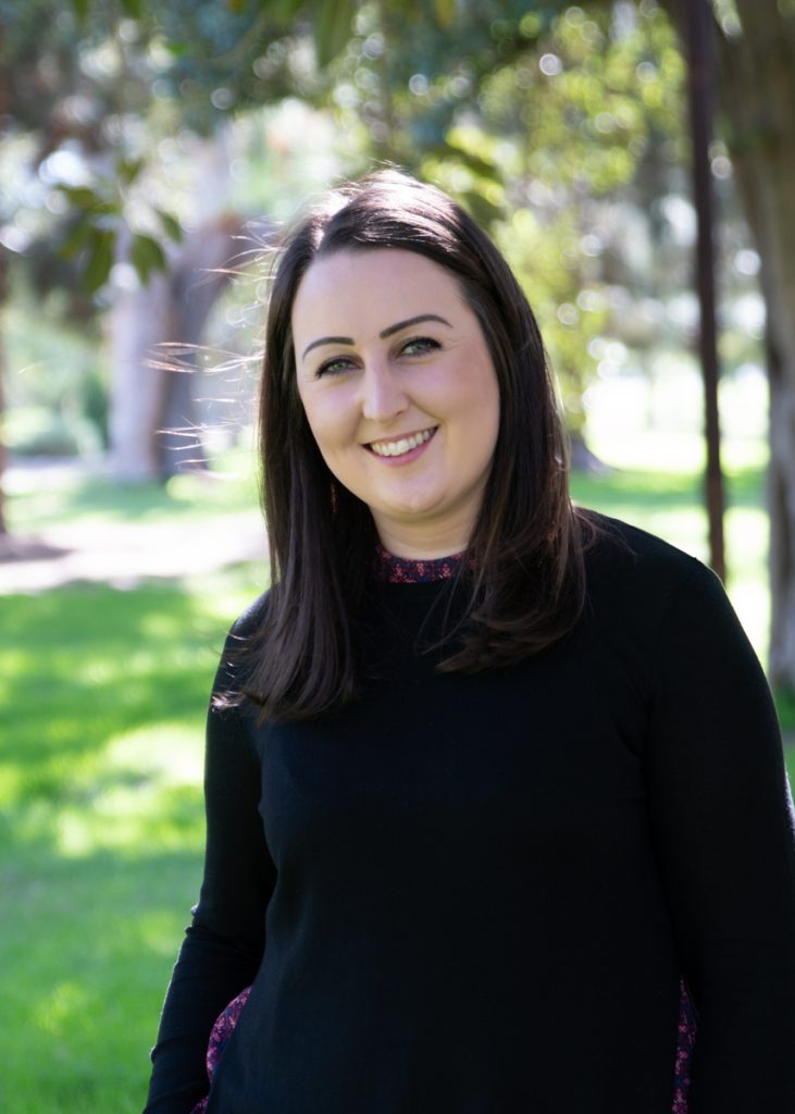 A woman with straight, dark brown hair smiles while standing outdoors. She is wearing a black long-sleeved top. The background is filled with green grass, trees, and dappled sunlight.