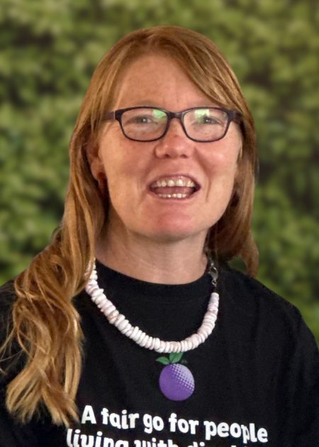 A woman with red hair and glasses smiles, wearing a white-beaded necklace and a black Purple Orange t-shirt, against a leafy green background.