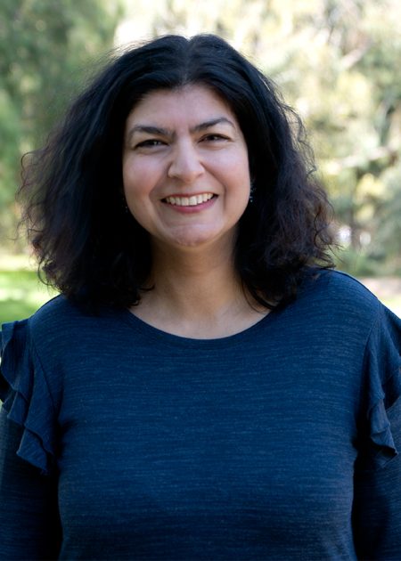 A woman with medium-length, curly dark hair smiles at the camera while standing outdoors. She is wearing a dark blue top with ruffled sleeves. The background shows a sunlit park with blurred trees and greenery.