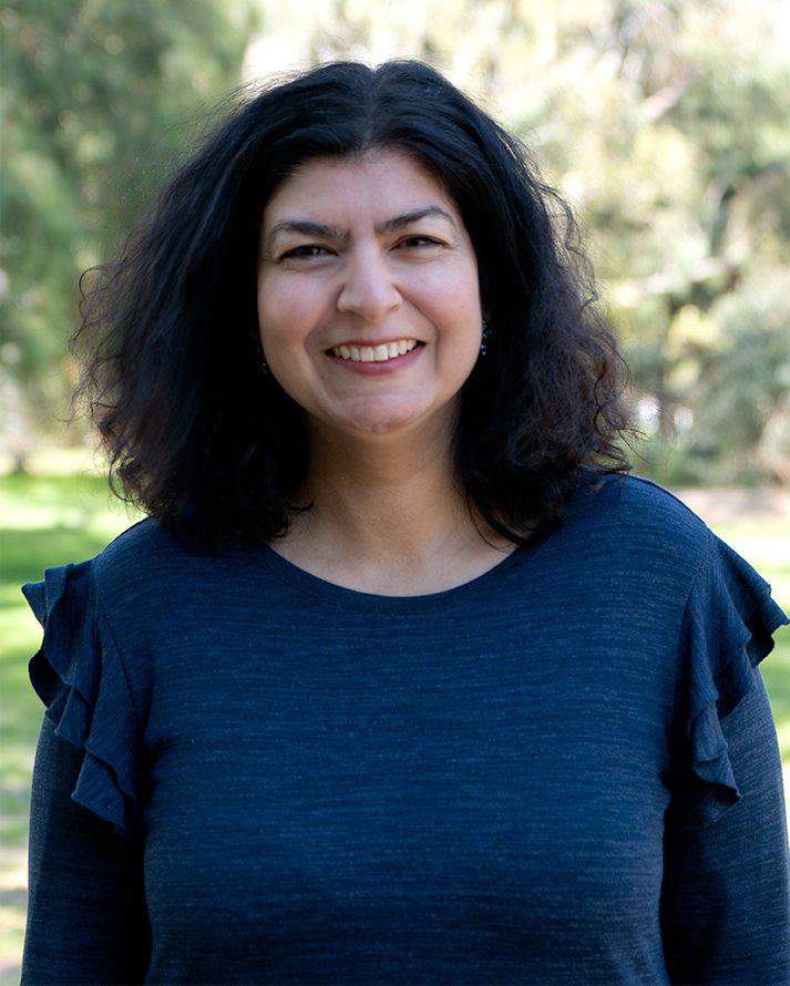 A woman with medium-length, curly dark hair smiles at the camera while standing outdoors. She is wearing a dark blue top with ruffled sleeves. The background shows a sunlit park with blurred trees and greenery.