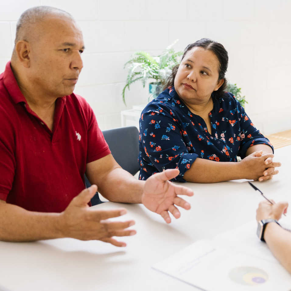 A man and a woman sitting at a table in an interview situation. The man on the left is bald and has a red t-shirt and the woman on his right, who has long dark hair, is looking in his direction.
