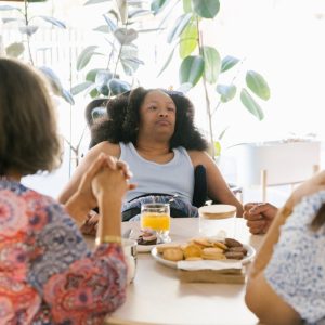 Young female wheelchair user seated at kitchen table. Two out of focus people are visible from behind. There are biscuits and a glass of juice on the table.