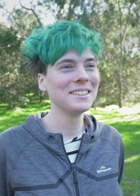 A person with bright green short hair smiles while standing outdoors. They are wearing a grey Kathmandu jacket over a black and white striped top. The background features trees and grass in soft focus.