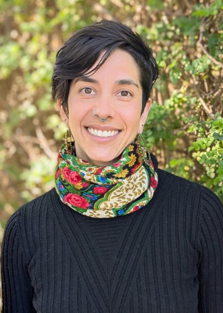 A person with short dark hair smiles warmly while standing in front of a leafy green background. They are wearing a black ribbed top and a colorful patterned scarf with floral and gold designs. They have a septum piercing and are wearing long, decorative earrings.