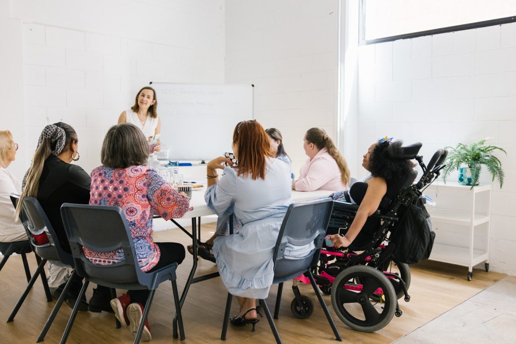 A group of people from different cultural backgrounds and with disability sit around a table looking at a presenter.