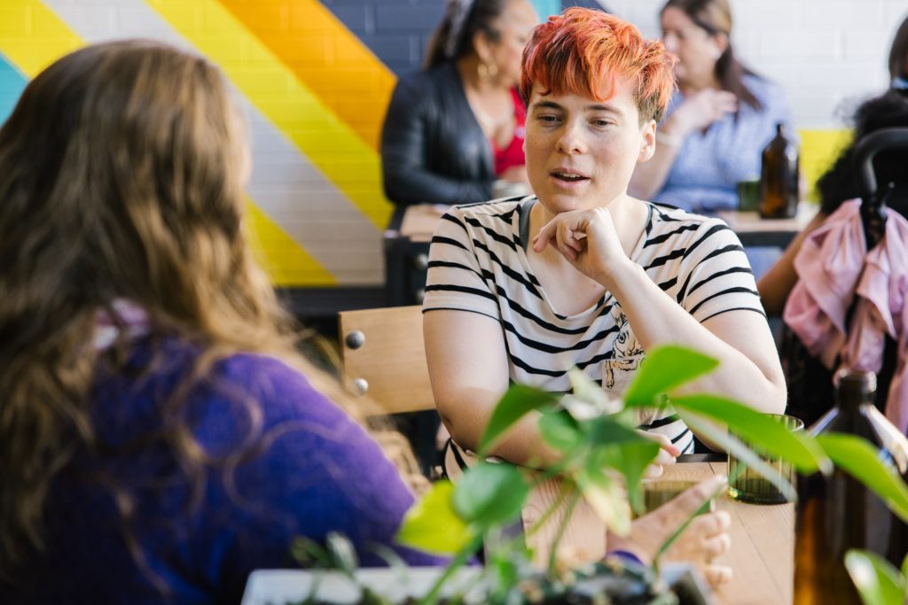 A person with short red hair and a striped top is talking to another person with long brown hair and a purple top. They are sitting at a cafe table.