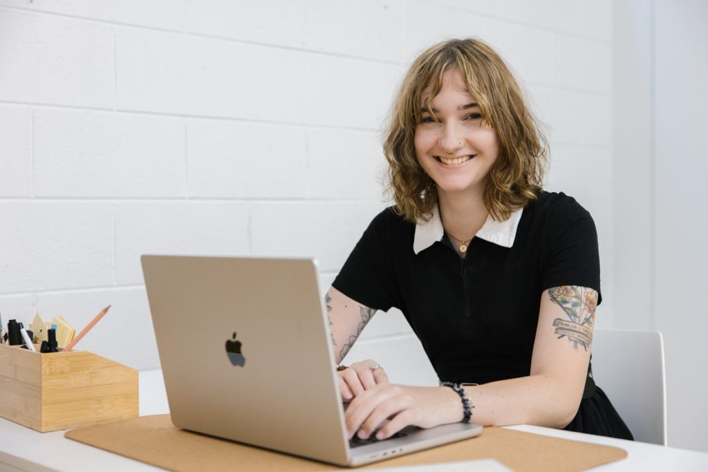 A person with medium length brown hair wearing a black top is typing on a laptop and smiling at the camera