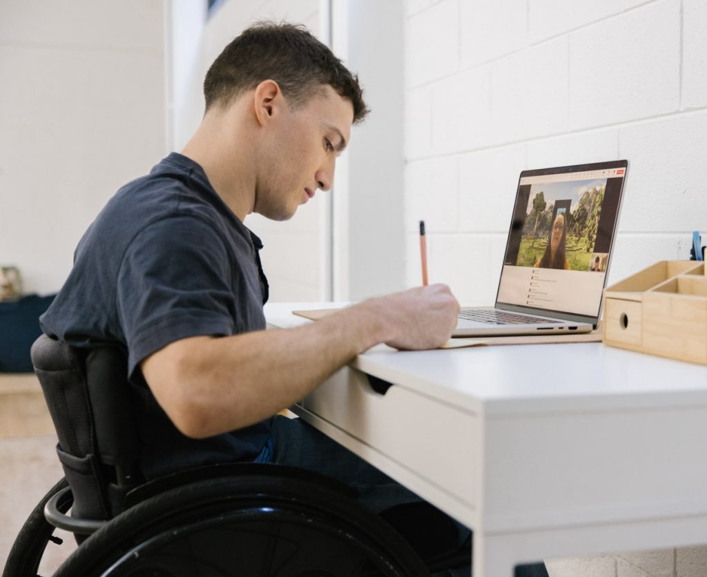 A man using a wheelchair sits at a white desk, writing in a notebook while participating in a video call on his laptop. A woman is visible on the screen with a nature background. The setting is a bright indoor space with a white wall and a wooden desk organiser.