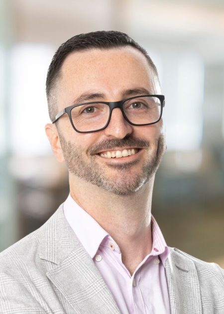 A man with short dark hair and glasses smiles confidently at the camera. He is wearing a light grey blazer over a pale pink shirt. The background is a softly blurred modern office space.