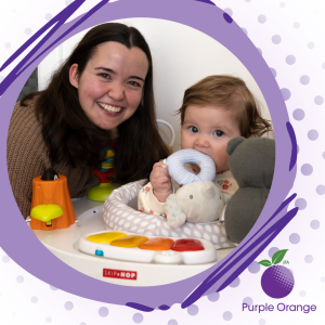 Light purple and spotty background. A grey purple misshapen circle with dark purple lines borders a photo of Lily and her baby. Lily smiles and they're both looking at the camera. They sit at a baby's play chair with a few toys around them