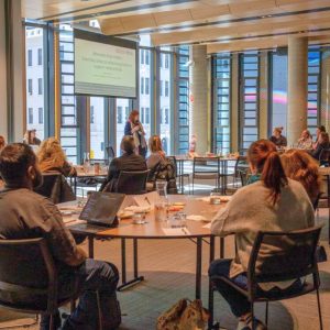 A group of people sit at round tables in a modern conference room with large windows, focused on a woman speaking at the front. A presentation slide behind her reads: "Moments that matter: Everyday steps to addressing harm in support relationships." Laptops, notepads, and water pitchers are on the tables, and the room is filled with natural light from the cityscape outside.