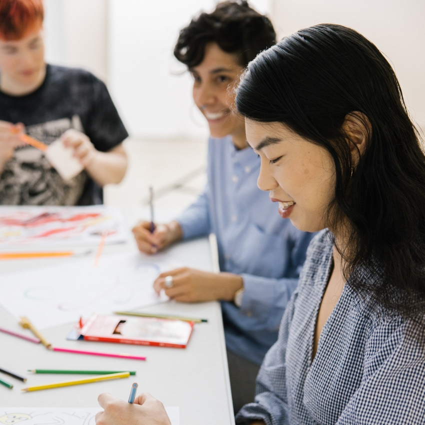 Three people sit together at a table, smiling and engaged in drawing or colouring in. The table is covered with art supplies and sheets of paper with sketches.