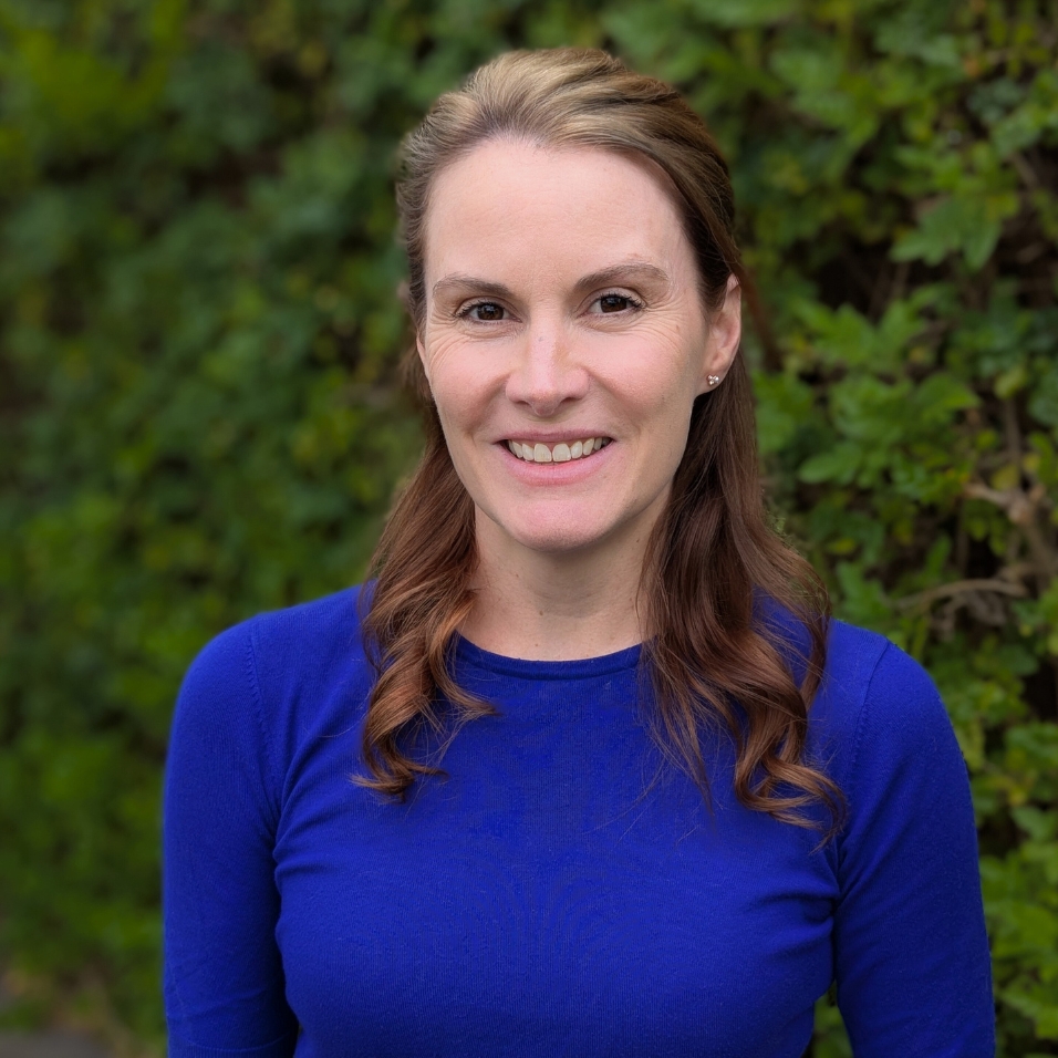 Smiling woman with long, wavy brown hair wearing a bright blue top, standing in front of a leafy green background.