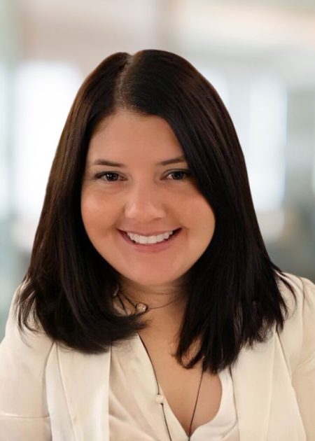 A woman with straight dark brown hair smiles warmly at the camera. She is wearing a white blazer and a light top, with a blurred professional indoor setting in the background.