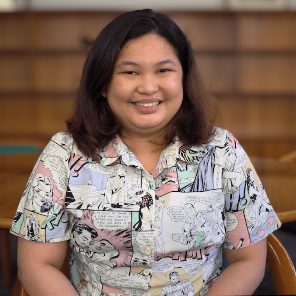 A person with shoulder-length dark hair smiles while seated indoors. They are wearing a colourful shirt featuring vintage-style comic book art. The background shows wooden shelves and tables, suggesting a library or study space.