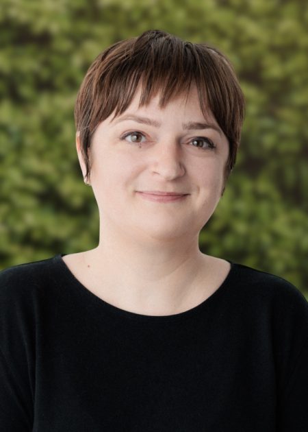 A person with short brown hair and a gentle smile stands in front of a leafy green background. They are wearing a plain black top and looking directly at the camera.