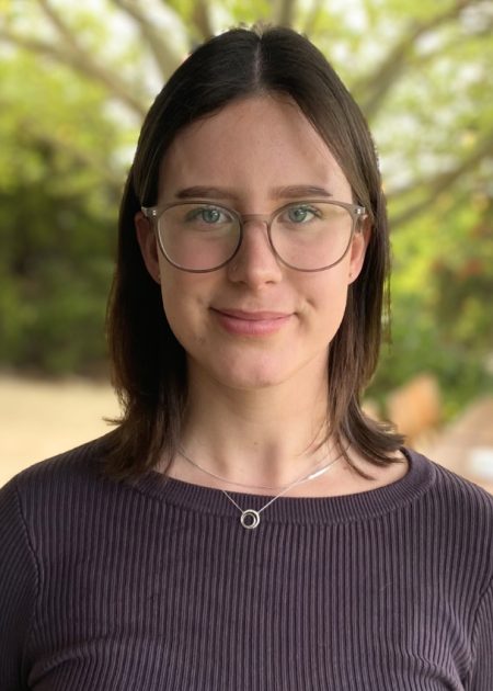 A woman with shoulder-length dark brown hair and clear-framed glasses smiles softly at the camera. She is wearing a ribbed, dark long-sleeve top and a silver necklace with a circular pendant. The background features a blurred outdoor setting with greenery and a long wooden table.