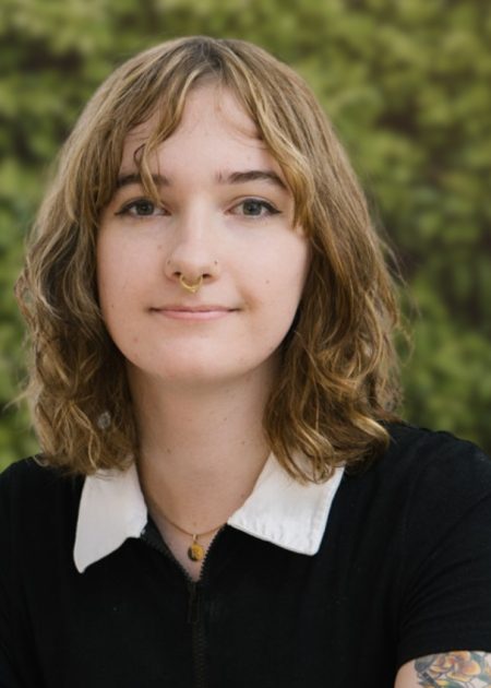 A person with wavy light-brown hair and a septum piercing looks at the camera with a slight smile. She wears a black top with a white collar and a small pendant necklace, with a tattoo visible on one arm. The background is a leafy green hedge in soft focus.