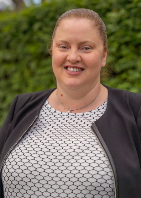 A person with light brown hair in a ponytail, wearing a geometric patterned white and black shirt and a black jacket. They are smiling at the camera.